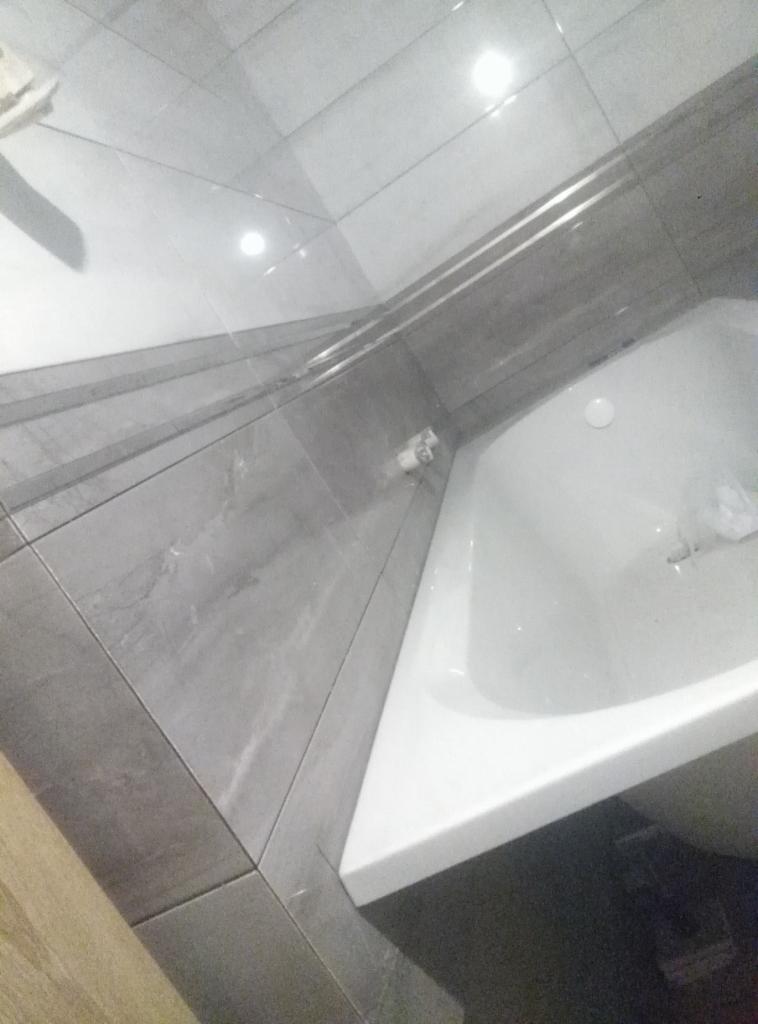 Narożna wanna w łazience z szarymi płytkami na ścianach i podłodze, widoczne elementy instalacji hydraulicznej przy wannie.