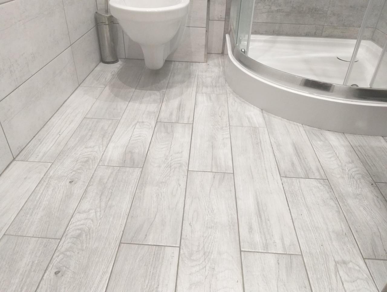 Jasnoszara łazienka z kabiną prysznicową i toaletą, podłoga wyłożona płytkami imitującymi drewno.