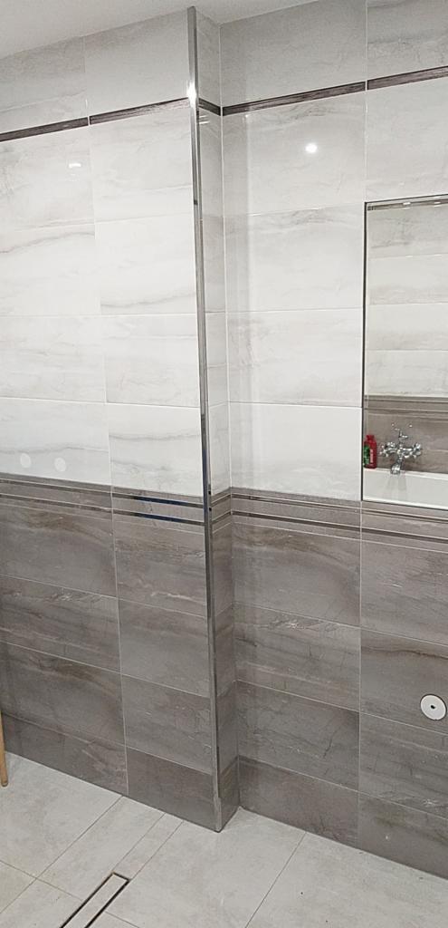 Wykończona łazienka z szarymi i białymi płytkami imitującymi marmur, z widocznym lustrem, baterią i odpływem liniowym w podłodze.