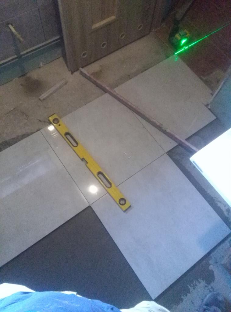 Układanie jasnych, kwadratowych płytek na podłodze, z użyciem poziomicy i lasera krzyżowego do precyzyjnego ustawienia, widoczny fragment kleju do płytek.