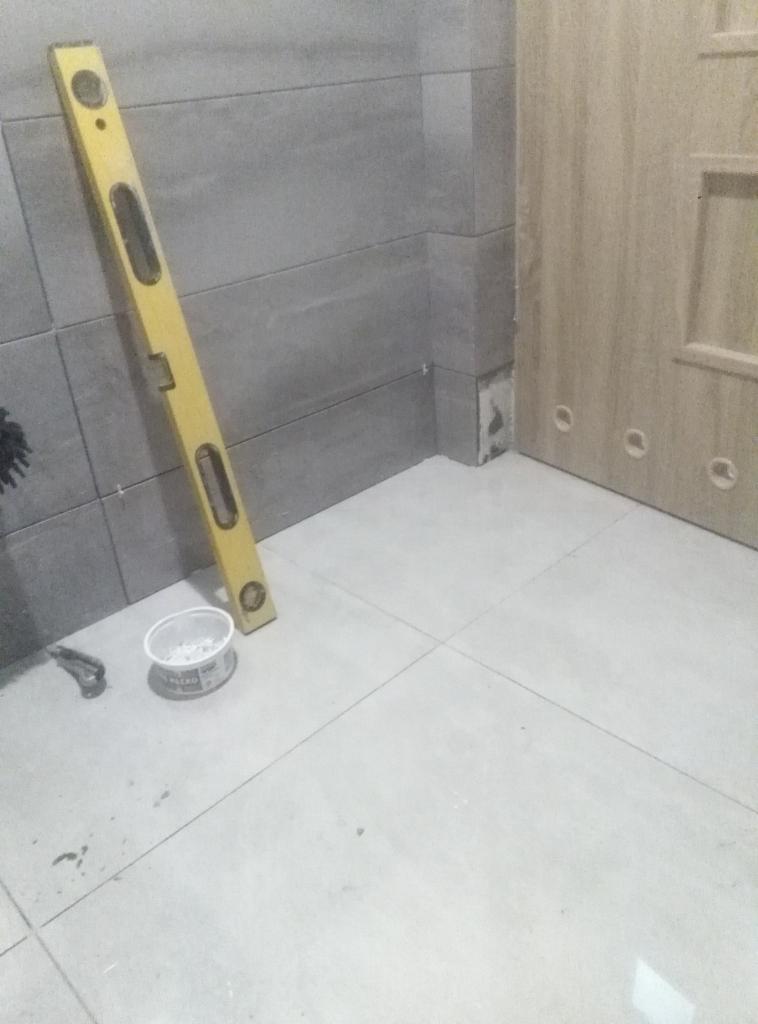 Ułożone szare płytki ścienne i białe płytki podłogowe w rogu łazienki, widoczna poziomica, pojemnik z zaprawą i szczypce.
