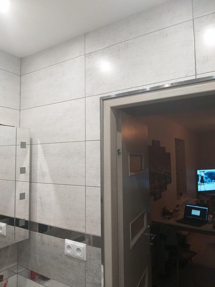 Wykończona łazienka z jasnymi, kwadratowymi płytkami imitującymi beton, z ciemniejszym pasem dekoracyjnym i białymi gniazdkami, widoczne obramowanie drzwi z metalową listwą i fragment pokoju...