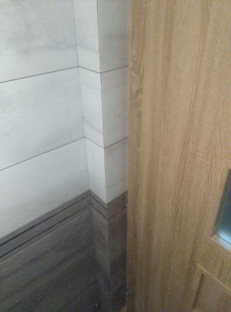 Wykończony narożnik łazienki z jasnymi płytkami ściennymi i ciemniejszymi płytkami podłogowymi imitującymi drewno, przylegający do drewnianej futryny drzwiowej.