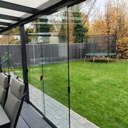 Remont Expert - Nowoczesna zabudowa tarasu z przesuwnymi szklanymi panelami, widok na zadbany trawnik z trampoliną w tle. Minimalistyczny design, szare płytki tarasowe i meble.