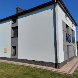 Dwukondygnacyjny dom z jasnoszarą elewacją, ciemnobrązowymi rynnami i oknami, z szarymi panelami dekoracyjnymi i wykończeniem z imitacji kamienia na fundamencie, otoczony trawnikiem.