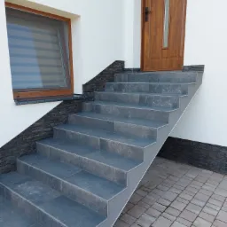 Zewnętrzne schody wejściowe wyłożone ciemnoszarą płytką z ryflowanymi krawędziami, prowadzące do drewnianych drzwi z pionowym przeszkleniem, obok okna z brązową ramą i żaluzjami, z elementami...