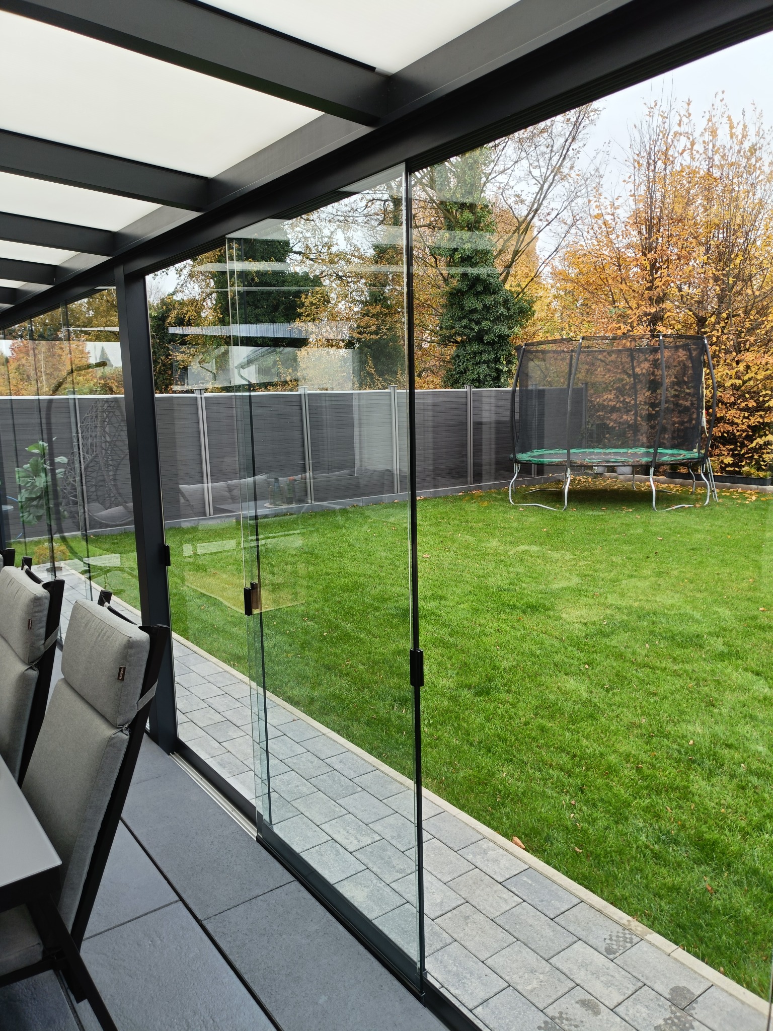 Nowoczesna zabudowa tarasu z przesuwnymi szklanymi panelami, widok na zadbany trawnik z trampoliną w tle. Minimalistyczny design, szare płytki tarasowe i meble.