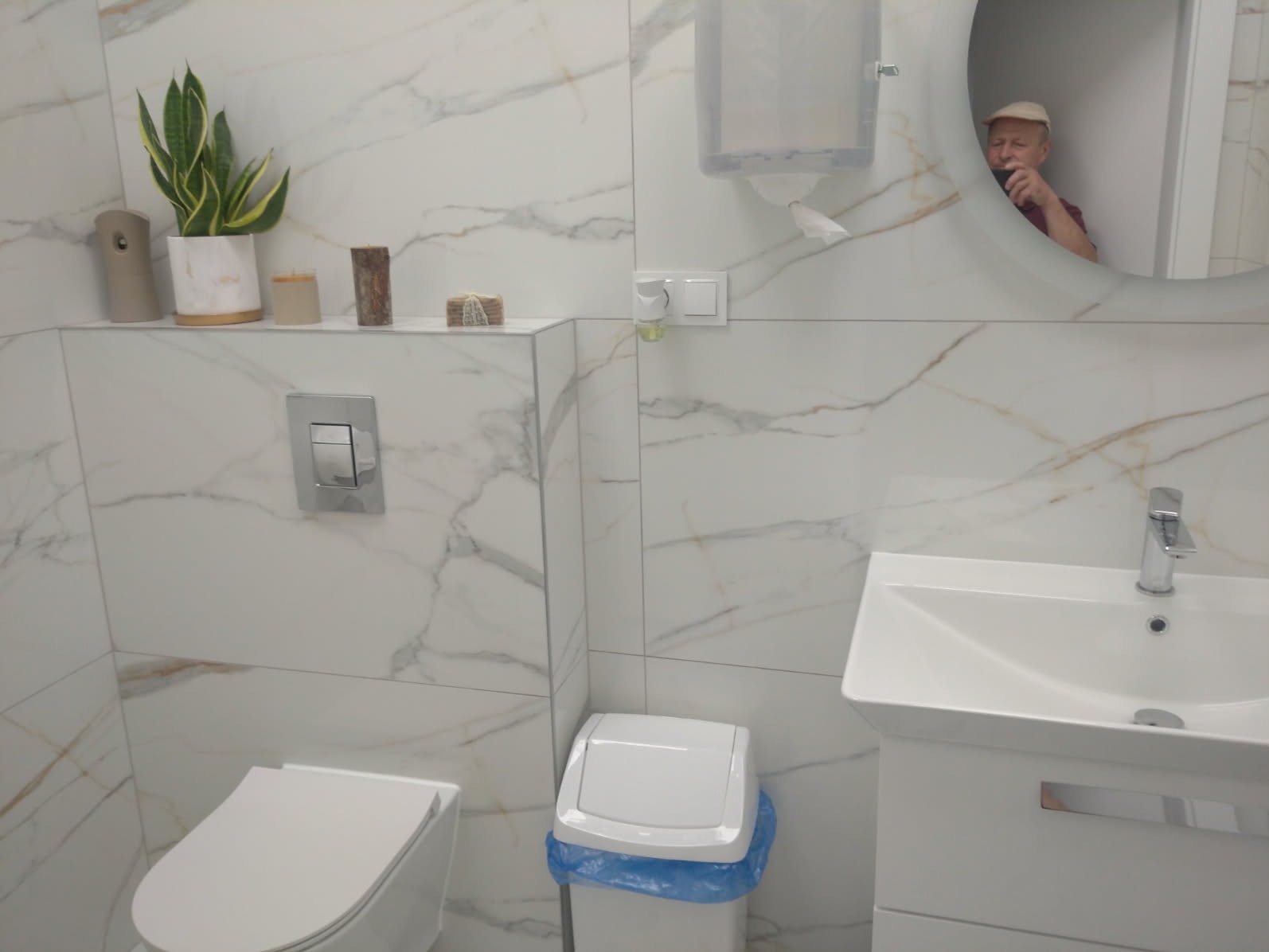 Biała, nowoczesna łazienka z marmurowymi płytkami, toaletą, umywalką i okrągłym lustrem, w którym odbija się osoba. Minimalistyczny design z roślinami na półce.