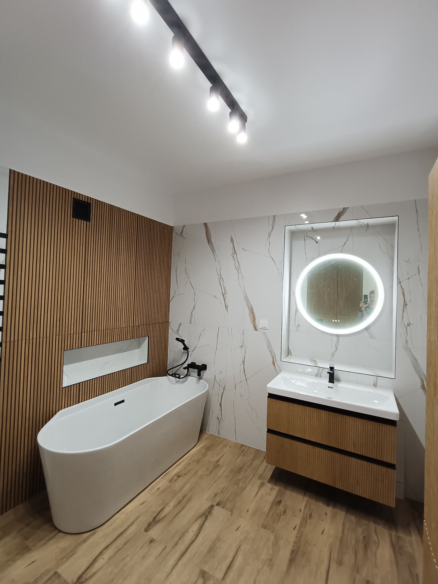 Nowoczesna łazienka z wanną, umywalką z szafką i okrągłym lustrem z podświetleniem LED. Ściany wykończone płytkami imitującymi marmur i drewnianymi lamelami.