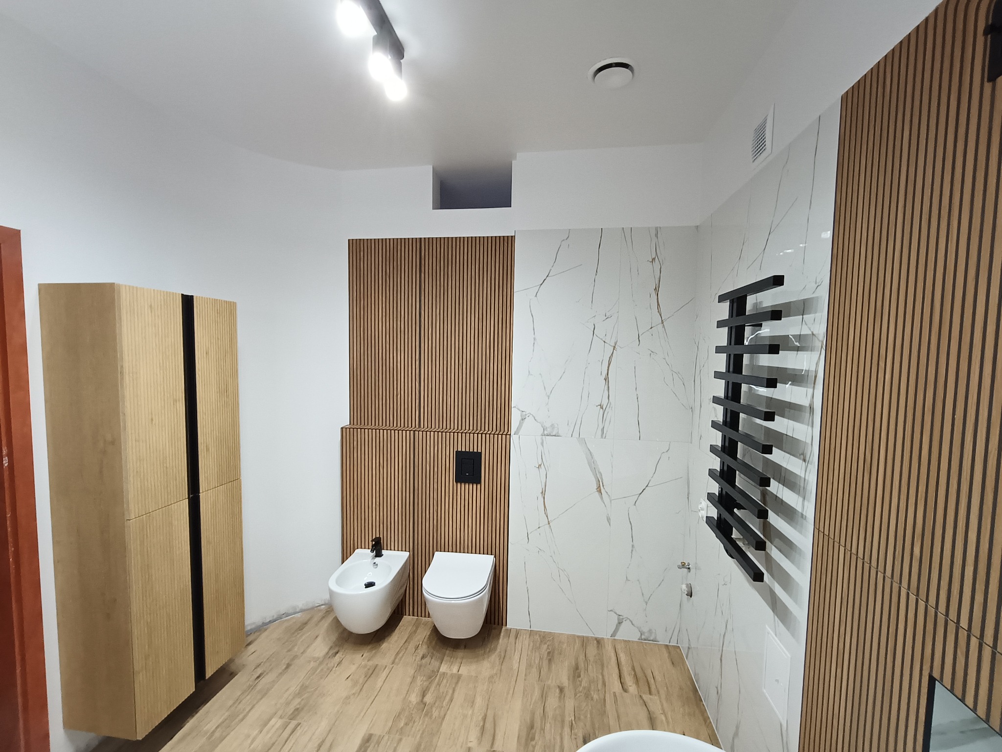 Nowoczesna łazienka po remoncie: białe ściany z marmurowym wzorem, drewniane panele, wisząca toaleta i bidet, czarny grzejnik drabinkowy, szafka z drewnianymi lamelami.