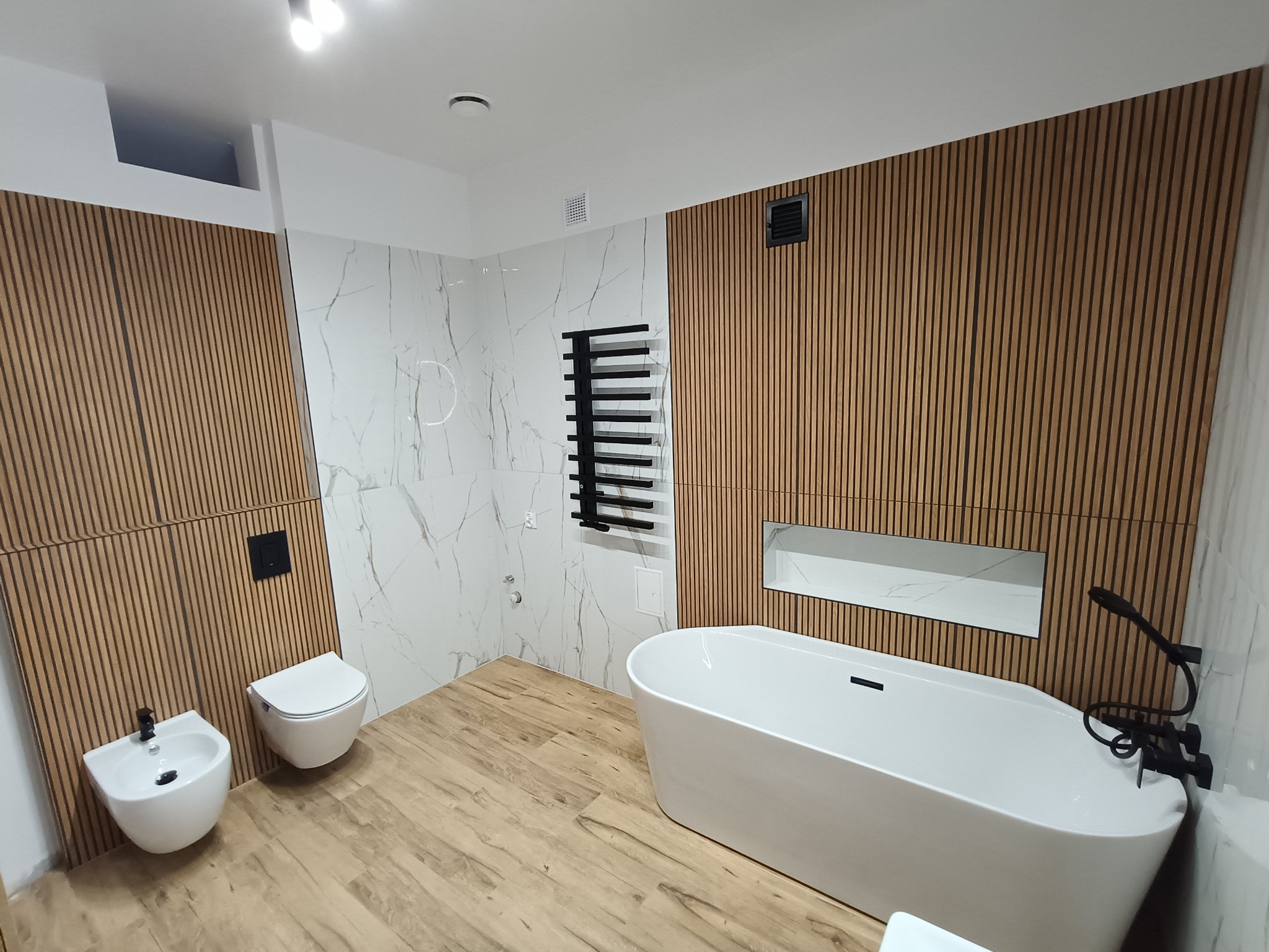Nowoczesna łazienka z drewnianymi panelami ściennymi, białą armaturą i marmurowymi płytkami. Minimalistyczny design z czarnymi akcentami.