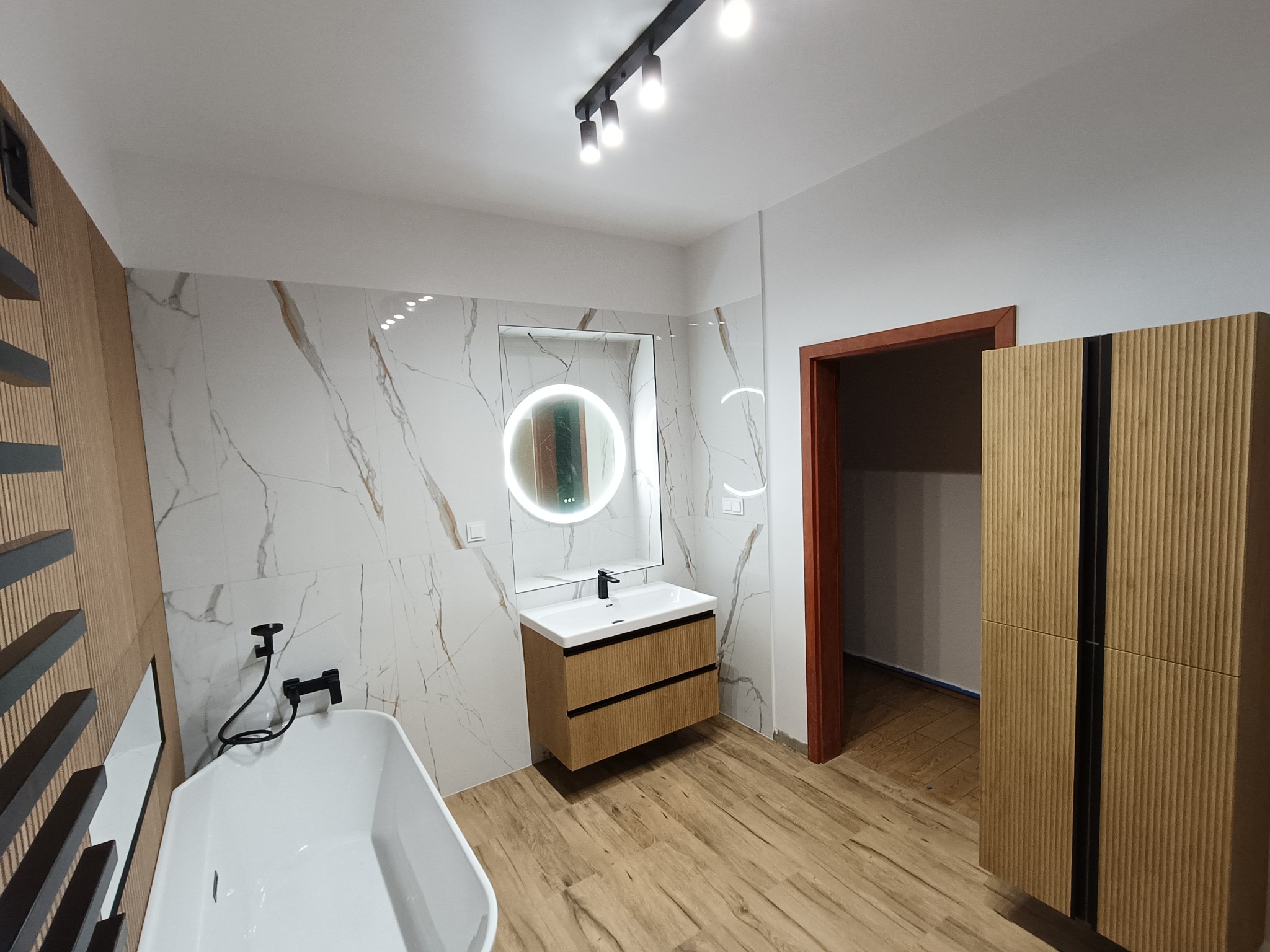 Nowoczesna łazienka z wanną, umywalką z szafką z lamelami, lustrem z podświetleniem LED i płytkami imitującymi marmur. Widoczne oświetlenie sufitowe i drewniana podłoga.