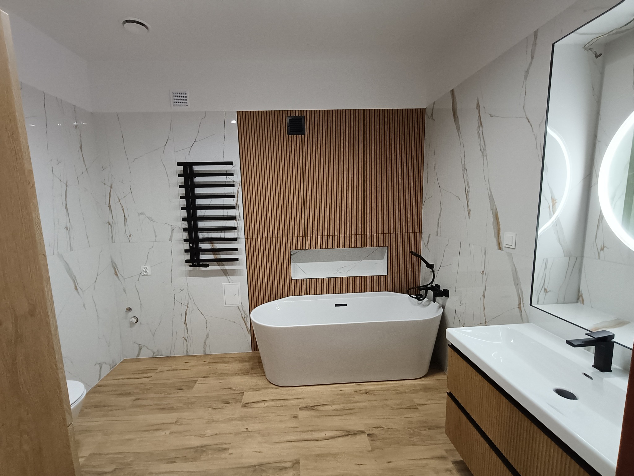 Nowoczesna łazienka z wolnostojącą wanną, drewnianą ścianą z lamelami i marmurowymi płytkami. Umywalka z szafką i okrągłe lustro z podświetleniem.