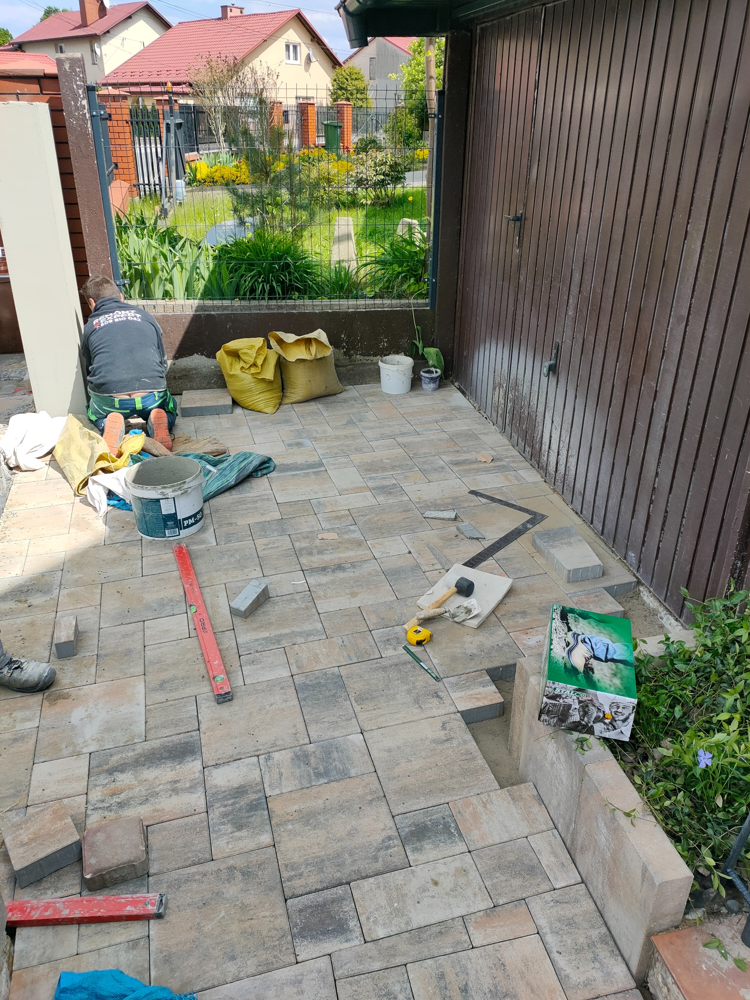 Układanie kostki brukowej na podwórku w Mielcu, widoczne narzędzia, poziomica, wiadro z zaprawą, częściowo ułożona nawierzchnia i pracownik w trakcie pracy.
