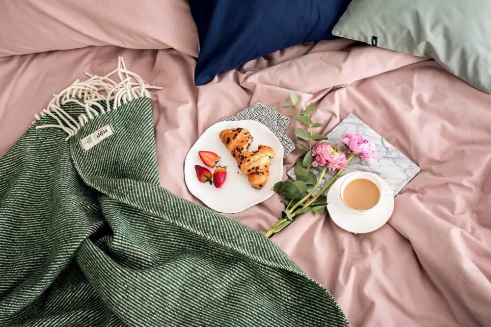 Jasnoróżowa pościel z zielonym pledem w jodełkę, croissantami z truskawkami, filiżanką kawy i kwiatami.