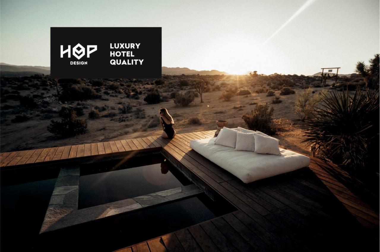 Luksusowy materac z poduszkami na drewnianym tarasie przy basenie, w tle pustynny krajobraz o zachodzie słońca, logo 'HOP DESIGN LUXURY HOTEL QUALITY' w lewym górnym rogu.