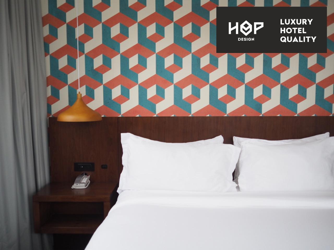 Hotelowe łóżko z białą pościelą, stolik nocny z telefonem i lampką, nad łóżkiem wzorzysta tapeta i logo 'HOP Design Luxury Hotel Quality'.