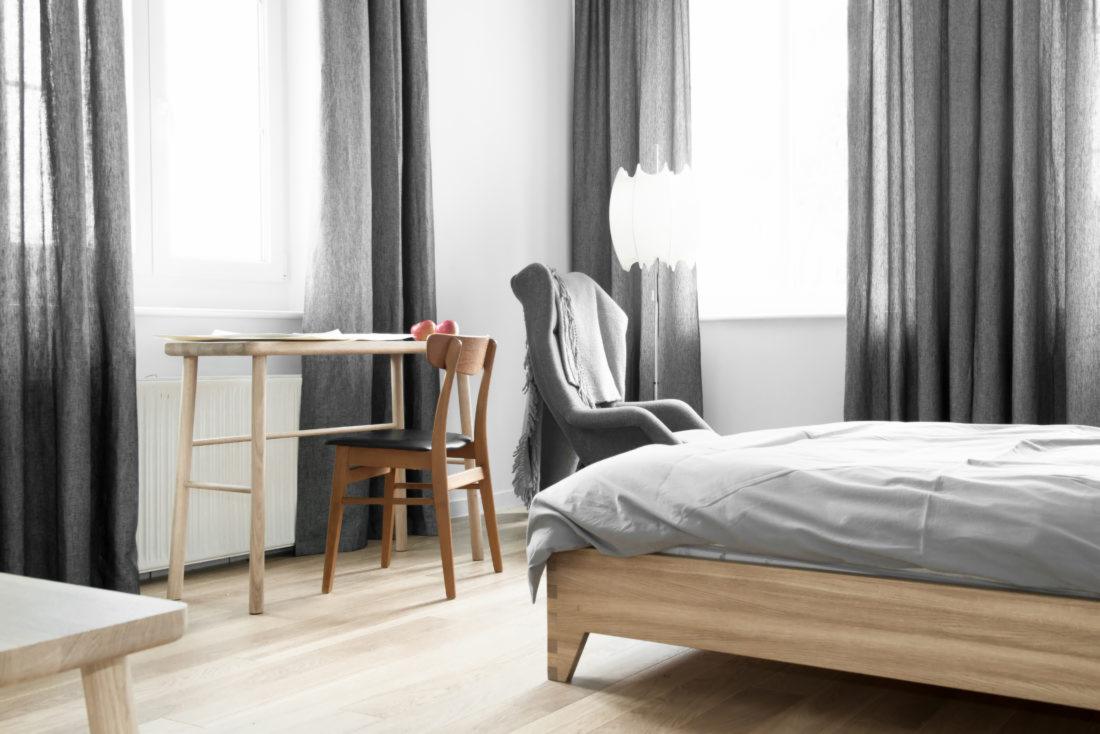 Jasne wnętrze sypialni z drewnianym łóżkiem przykrytym szarą pościelą, biurkiem z krzesłem oraz szarymi zasłonami w oknach. Na biurku leżą jabłka.