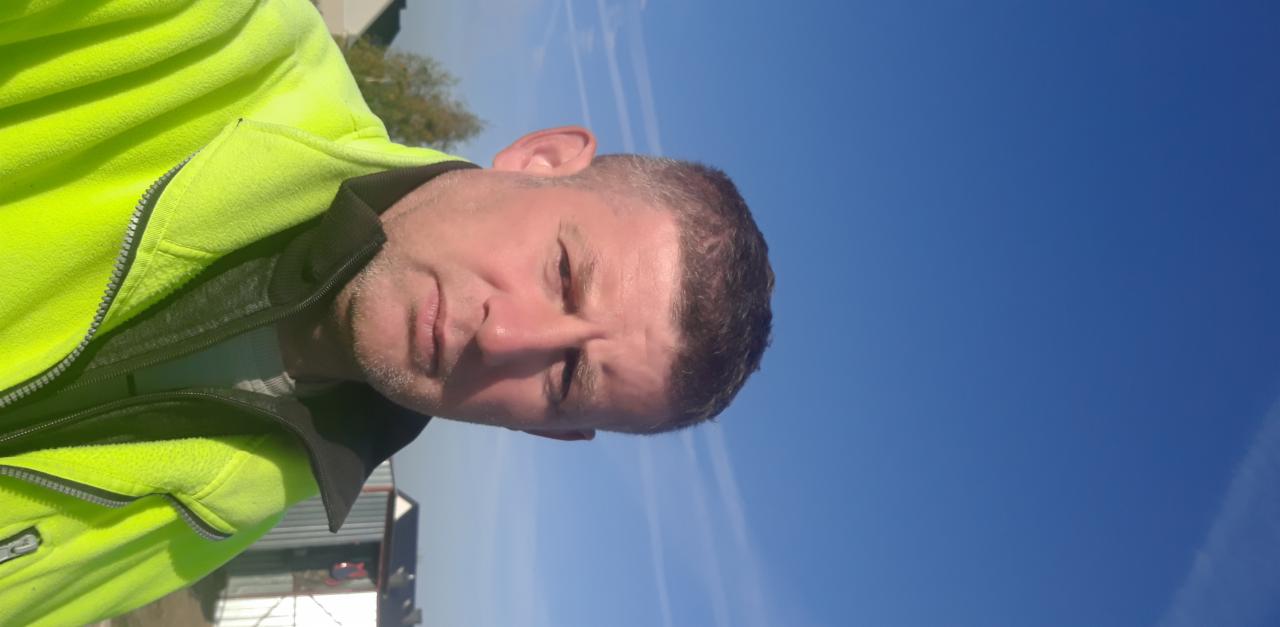 Mężczyzna w neonowej kurtce roboczej, z krótką fryzurą, na tle błękitnego nieba. Ujęcie z dołu, widoczny fragment budynku i drzewa w tle, obrócone o 90 stopni.