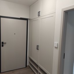 Stach Group Hubert Stachowski - Biała szafa wnękowa z czarnymi uchwytami, w przedpokoju obok drzwi wejściowych z czarną ramą. Widoczne półki na buty i regulator temperatury na ścianie.