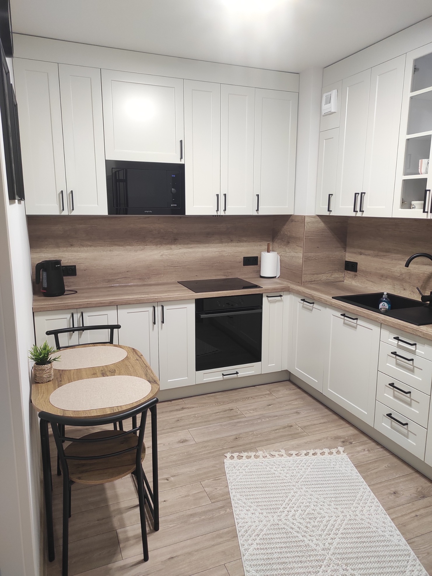 Jasna kuchnia z białymi szafkami i drewnianym blatem. Na blacie czajnik, mikrofalówka i płyta indukcyjna. Stół z krzesłami i dywanik na podłodze.