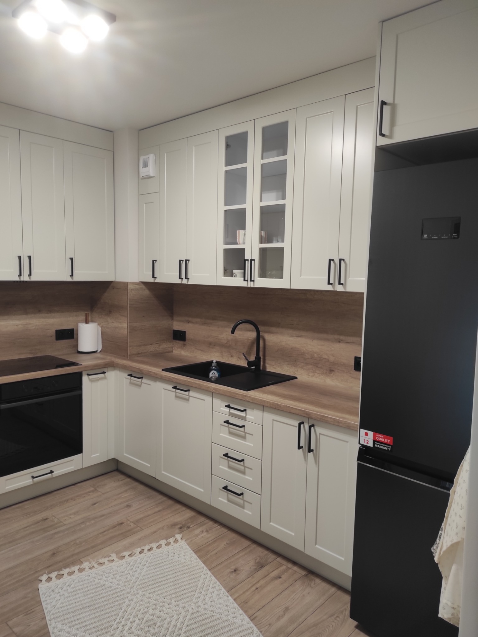 Jasna kuchnia z drewnianym blatem i czarnymi akcentami: bateria, zlew, uchwyty. Na podłodze dywanik. Widoczna lodówka. Nowoczesny design.