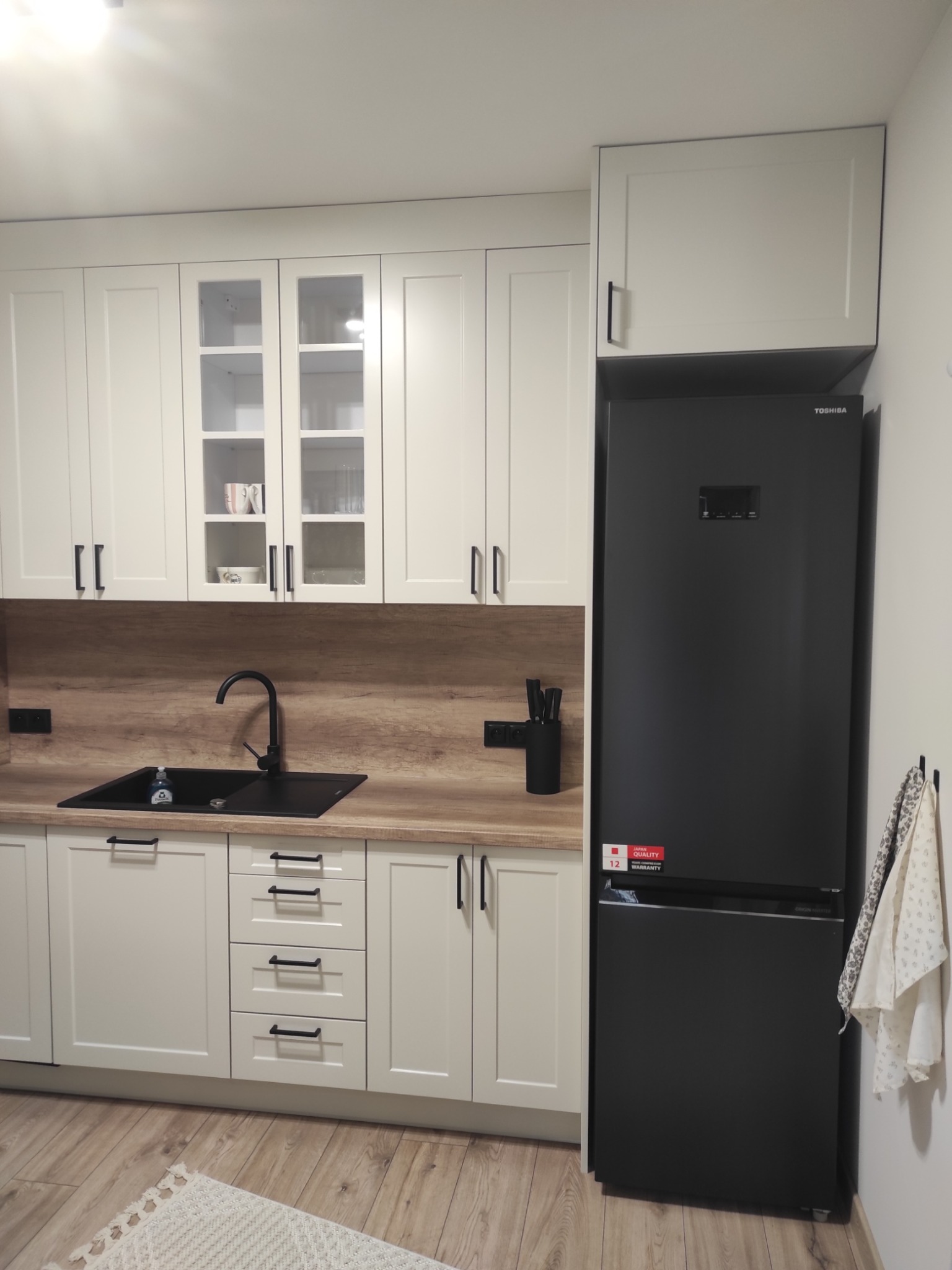 Jasna kuchnia z czarnymi akcentami w Osiecznej: zabudowa z frezowanymi frontami, czarny zlew i bateria, lodówka Toshiba. Nowoczesny design.