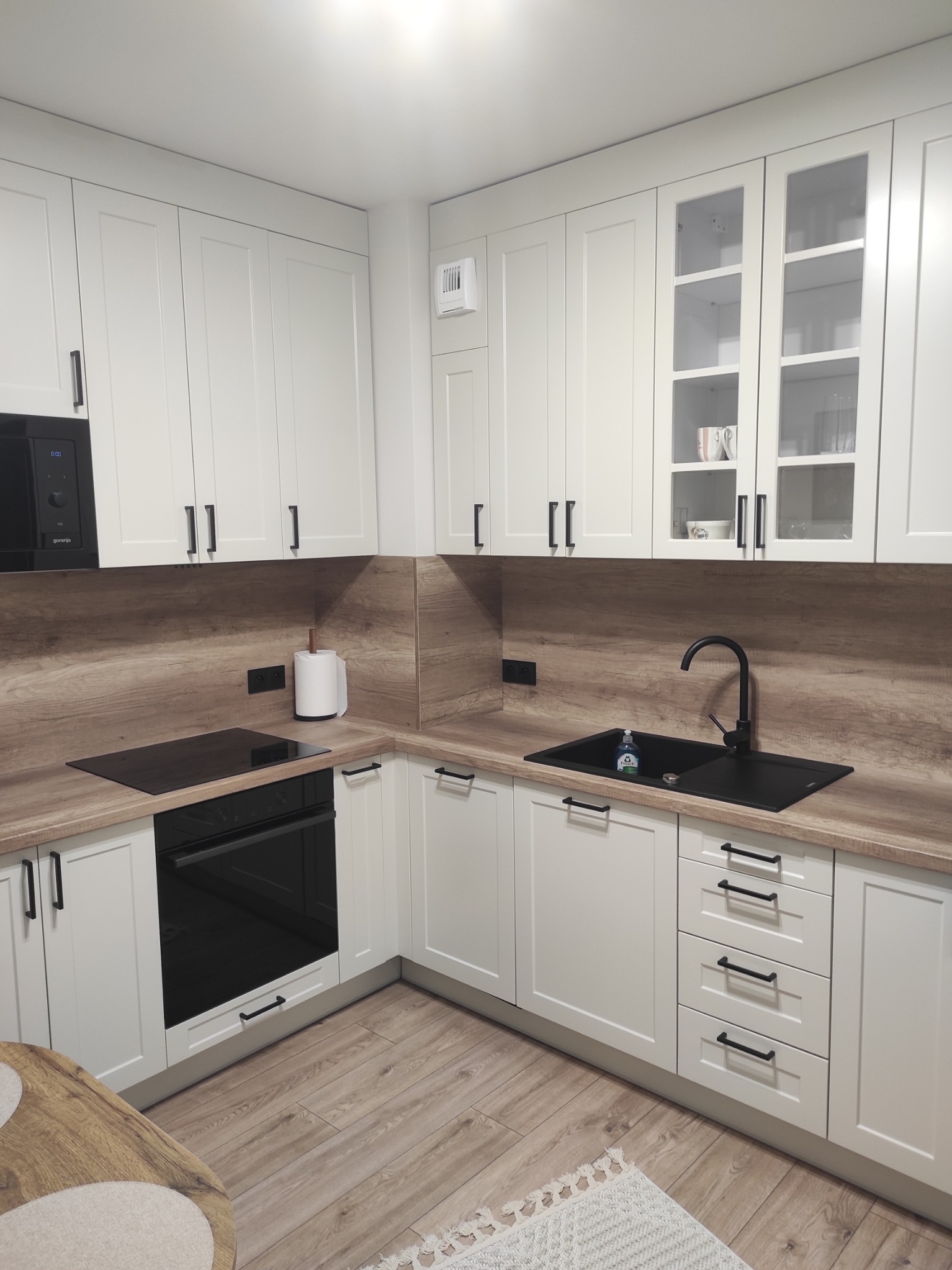 Jasna kuchnia w kształcie litery L z drewnianym blatem i czarnymi akcentami: zlew, bateria, uchwyty. Na blacie kuchennym widoczny dozownik do mydła.