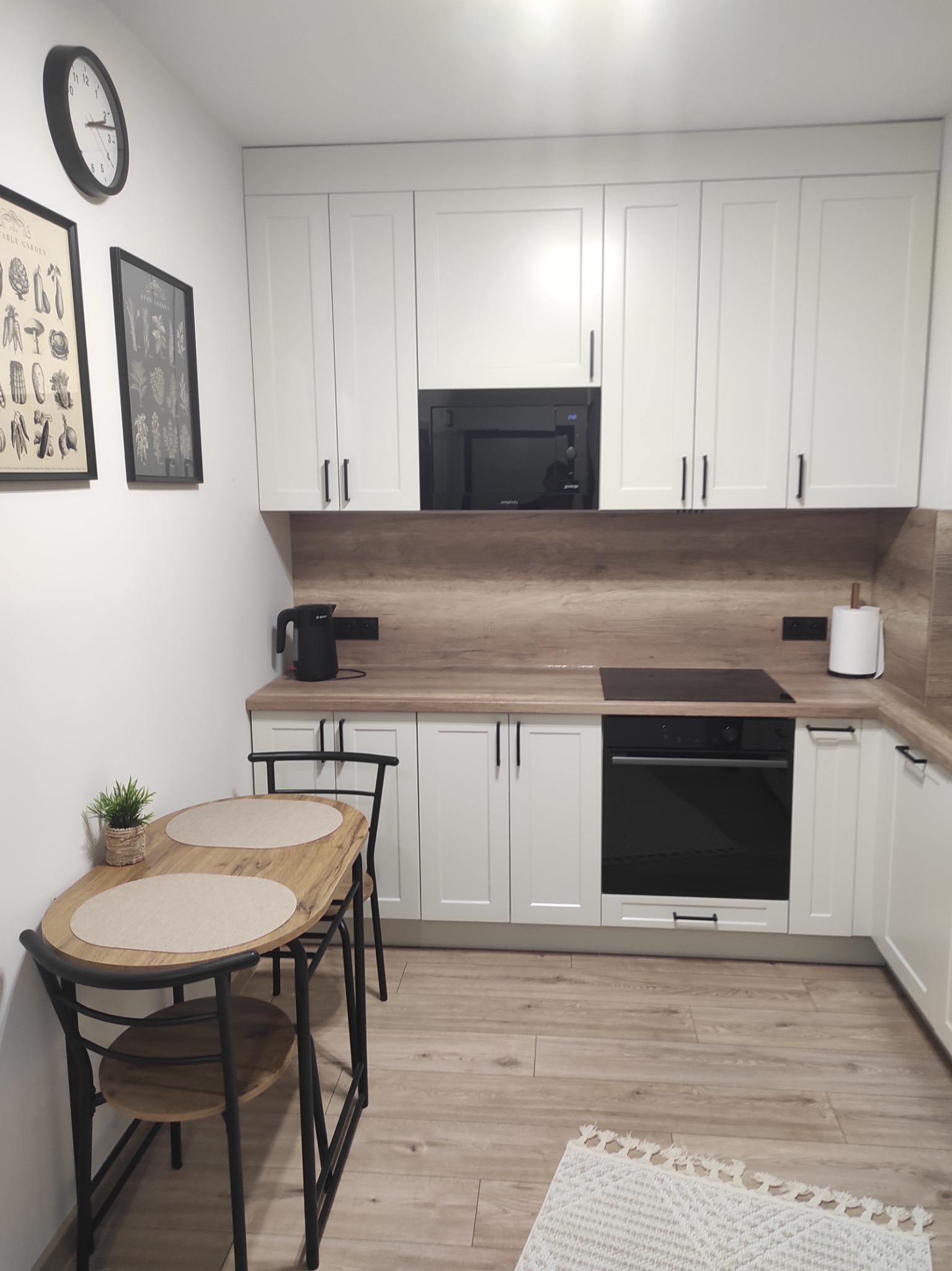 Biała kuchnia z drewnianym blatem i czarnymi akcentami: piekarnik, mikrofalówka, armatura. Stół z krzesłami i dekoracje na ścianie dopełniają aranżację.