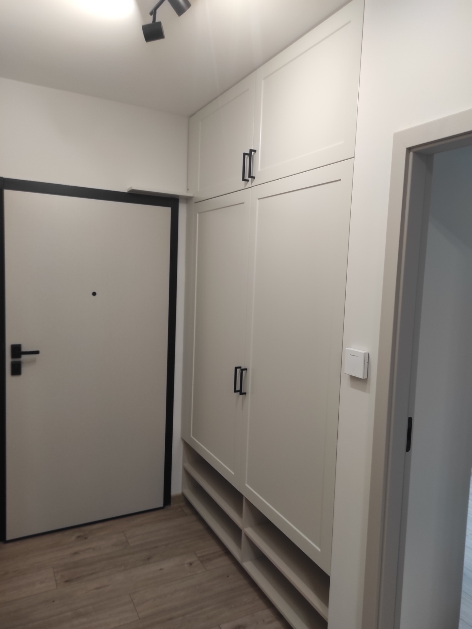 Biała szafa wnękowa z czarnymi uchwytami, w przedpokoju obok drzwi wejściowych z czarną ramą. Widoczne półki na buty i regulator temperatury na ścianie.