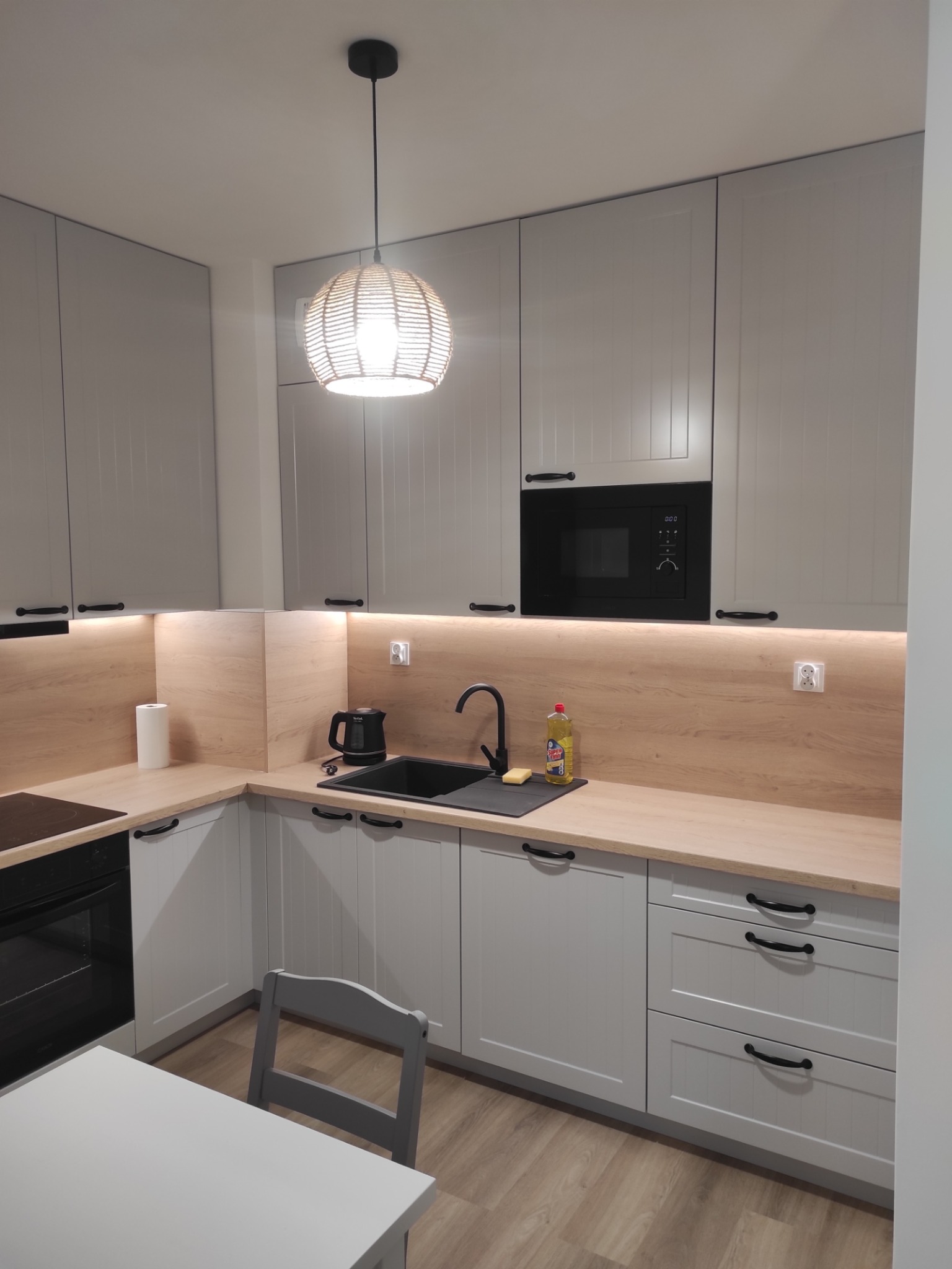 Jasnoszara kuchnia z drewnianym blatem i czarnymi uchwytami. Nad zlewem wisi okrągła lampa z wikliny. Widoczny czajnik i mikrofalówka.