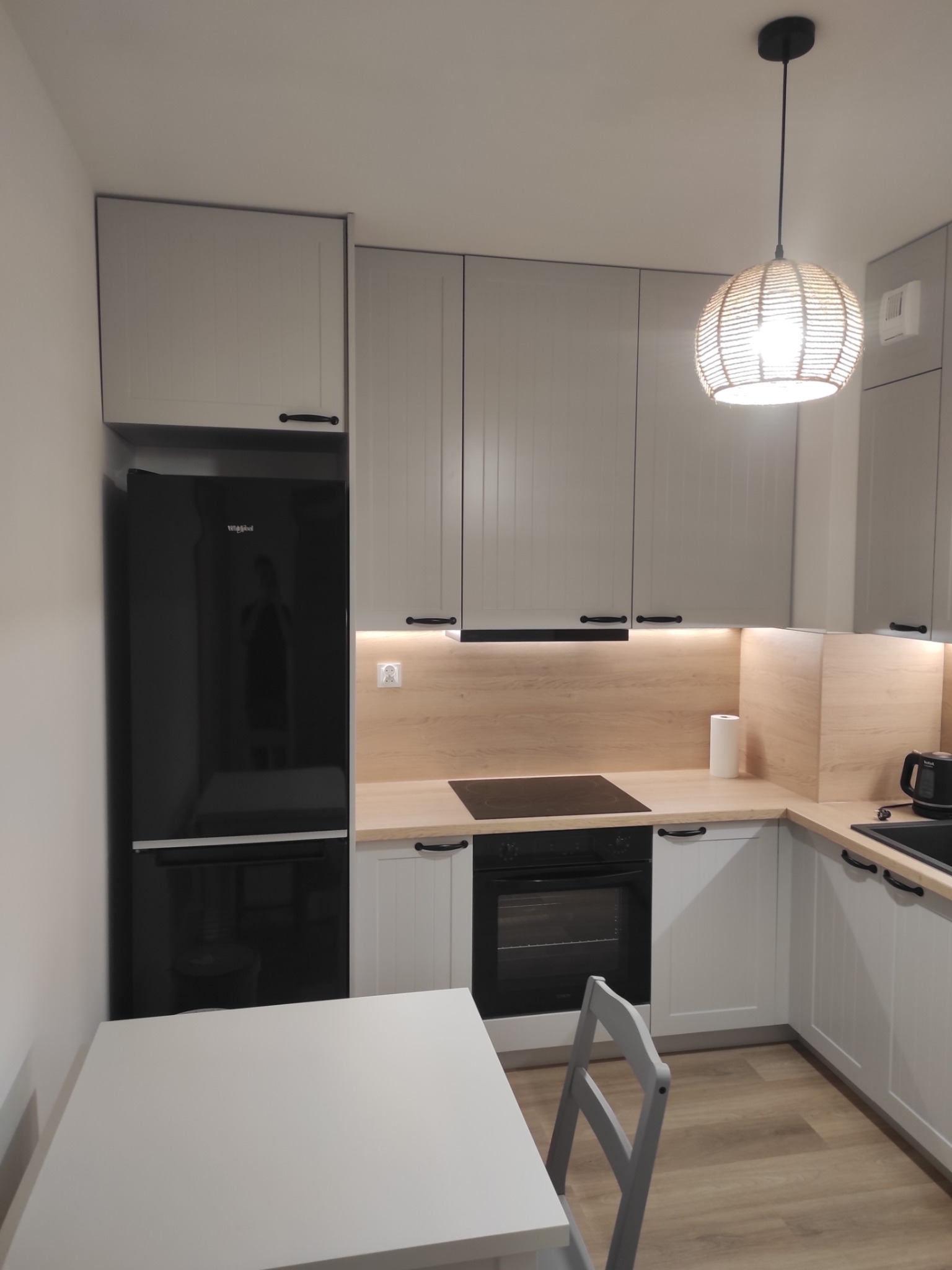 Jasnoszara kuchnia na wymiar z czarnym sprzętem AGD i drewnianym blatem. Nad blatem oświetlenie LED. W tle widoczny stół i krzesło.