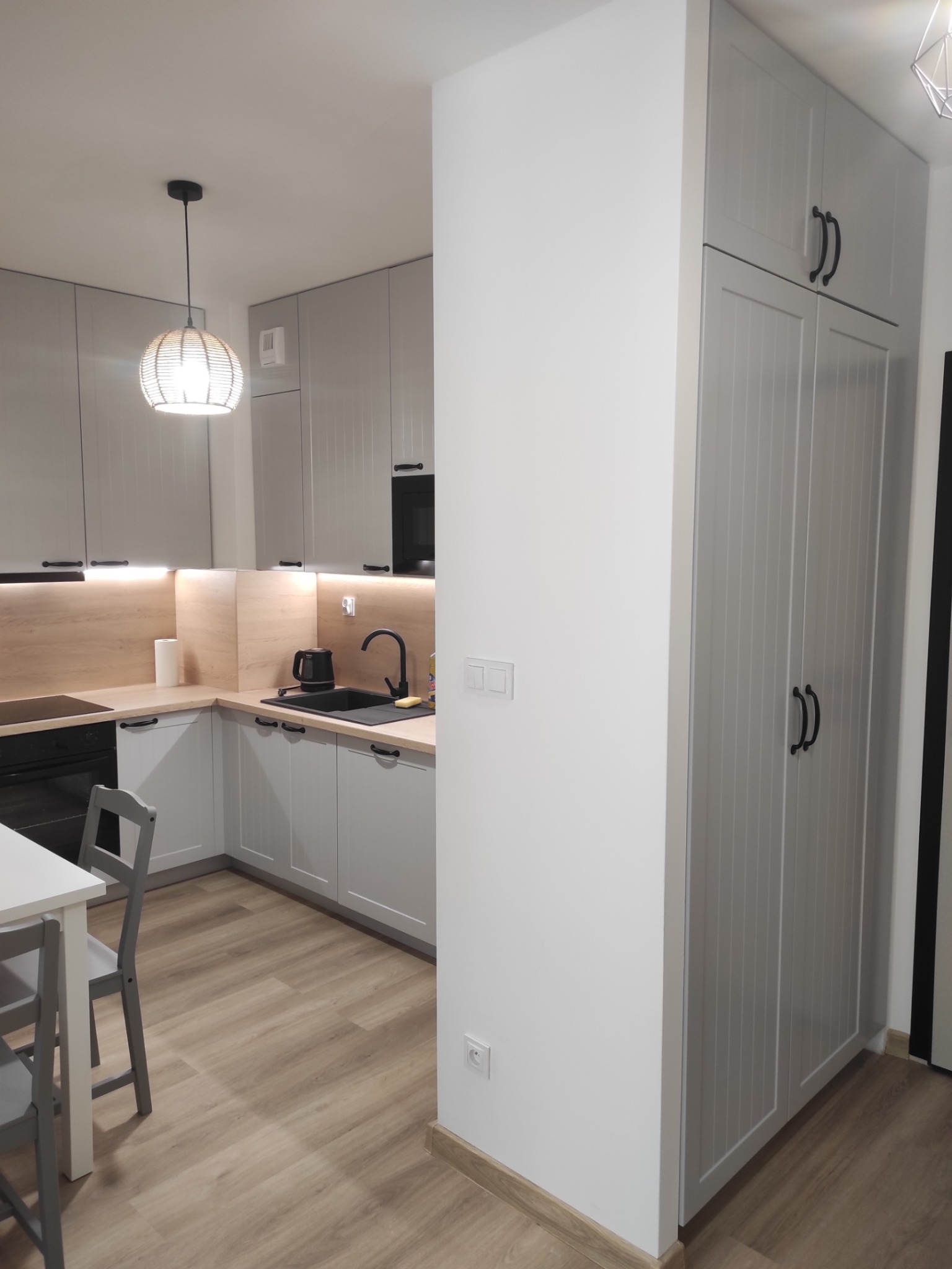 Jasnoszara kuchnia z drewnianym blatem i oświetleniem LED pod szafkami. Na pierwszym planie wysoka szafa z frezowanymi drzwiami i czarnymi uchwytami.
