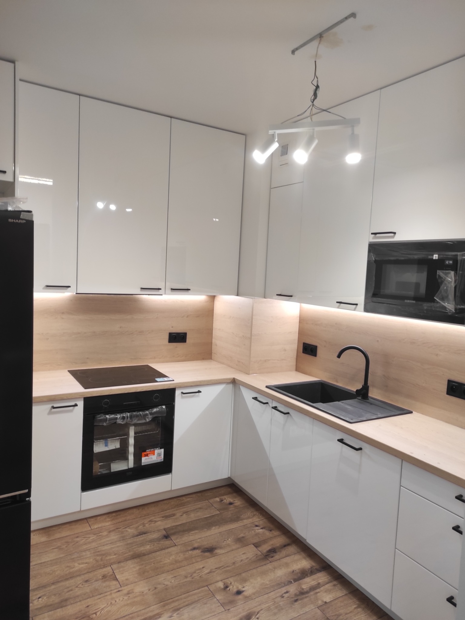 Biała kuchnia z drewnianym blatem i czarnymi akcentami: zlew, bateria, uchwyty. Podświetlenie LED pod szafkami. Widoczny piekarnik i płyta indukcyjna. Nowoczesny design.