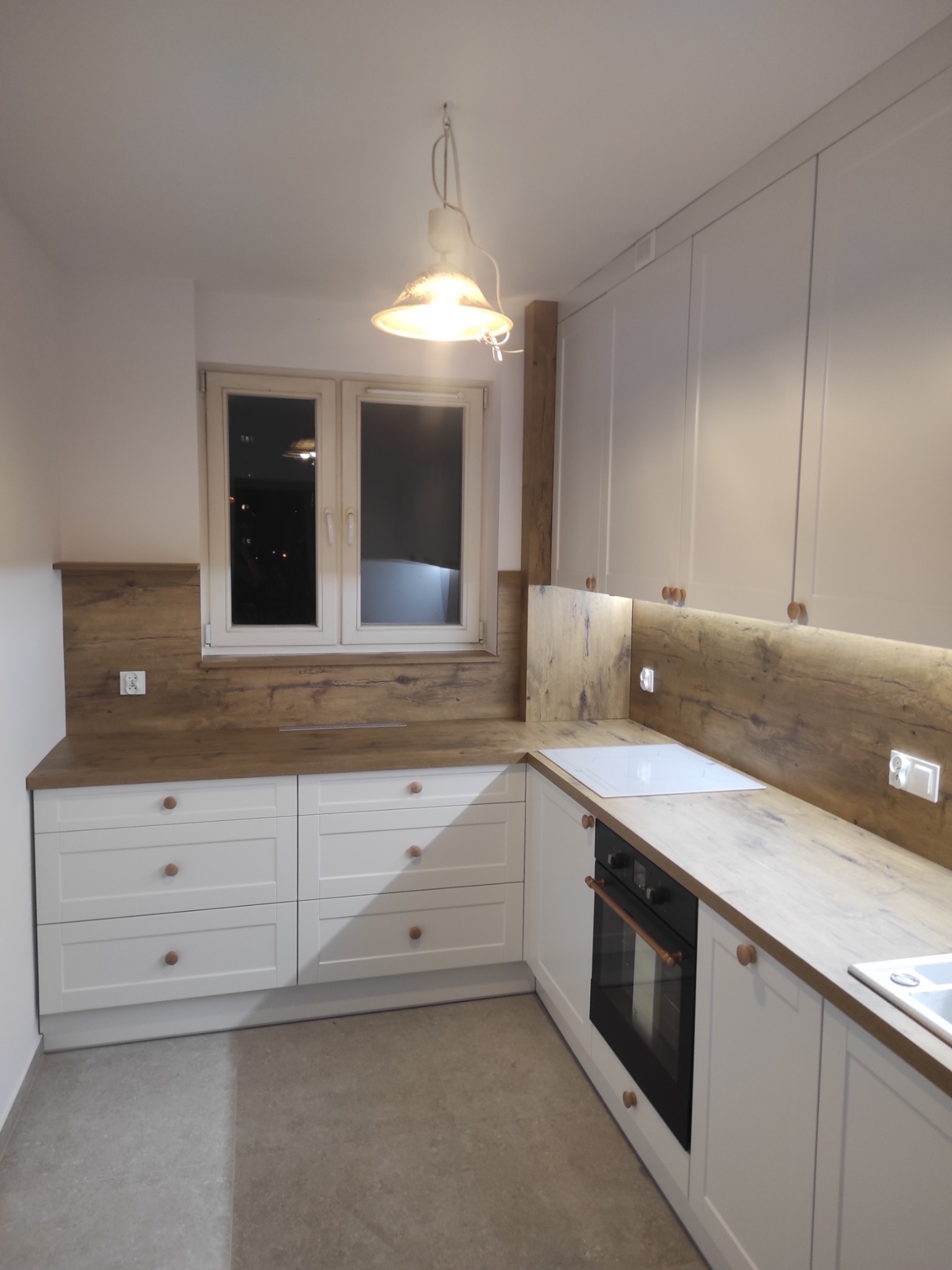 Jasna kuchnia z białymi szafkami i drewnianym blatem. Nad blatem zamontowane oświetlenie LED. Widoczne okno z widokiem na nocne miasto. Uwagę przykuwają okrągłe, drewniane uchwyty.