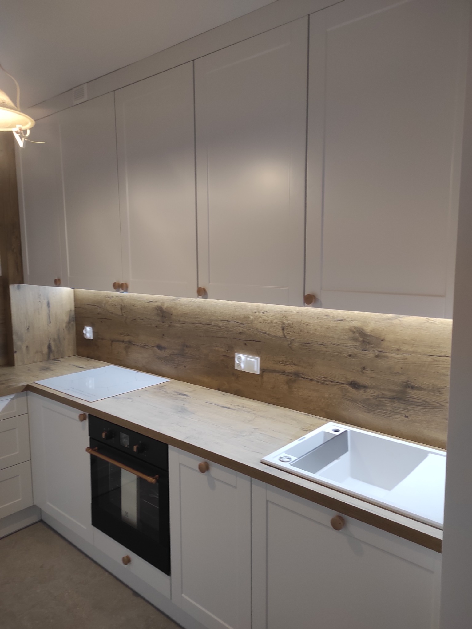 Jasna kuchnia z drewnianym blatem i białymi szafkami. Uwagę przykuwają okrągłe, drewniane uchwyty i podświetlenie blatu. Widoczna płyta indukcyjna i zlew.