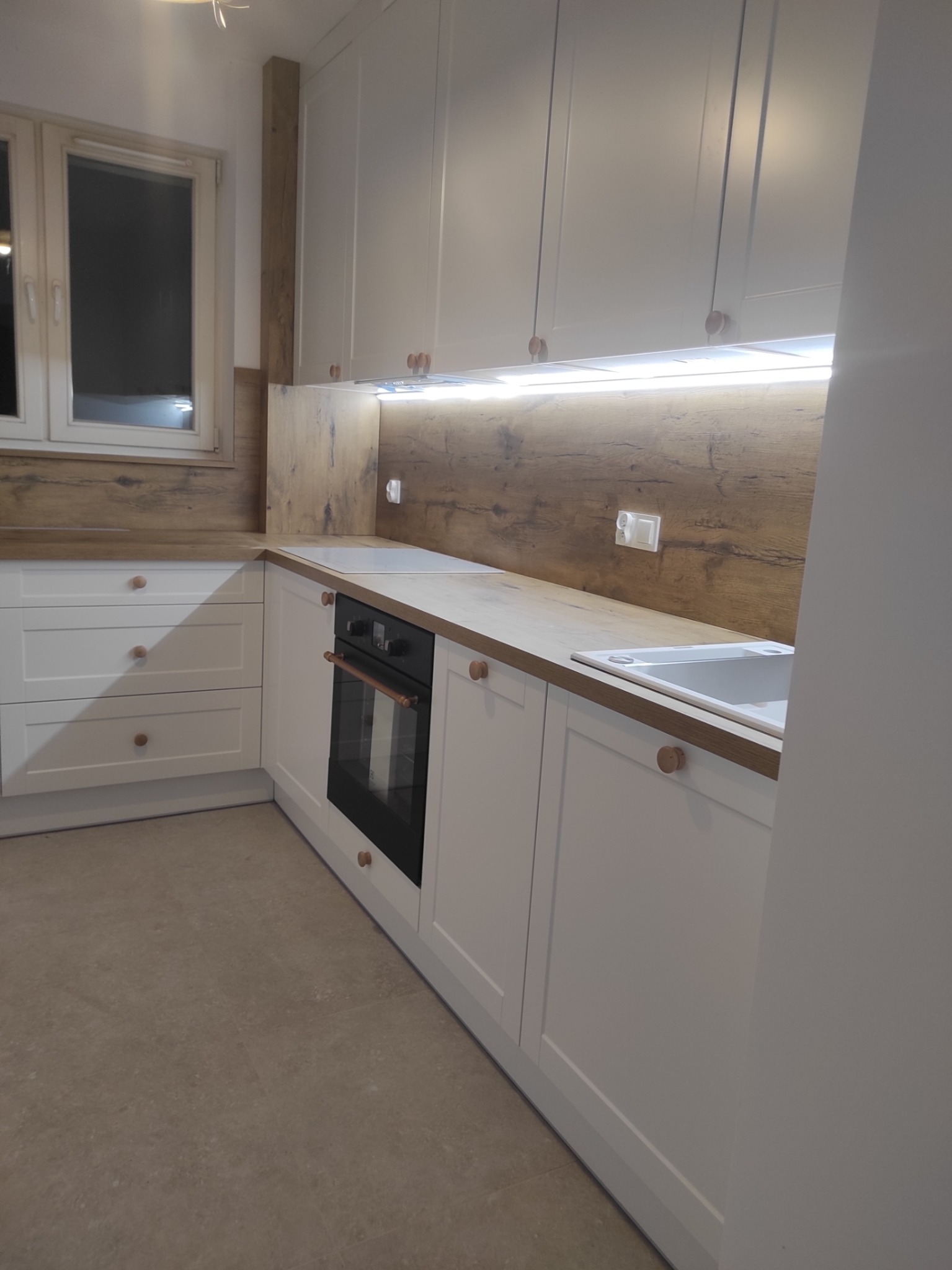 Jasna kuchnia z drewnianym blatem i oświetleniem podszafkowym. Widoczny piekarnik i zlew. Uwagę zwracają okrągłe, drewniane uchwyty.