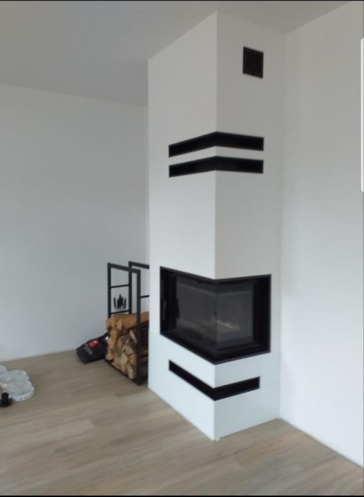 Biały narożny kominek z czarnymi akcentami w nowoczesnym wnętrzu, obok metalowy stojak z drewnem opałowym.