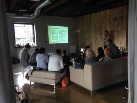 Grupa osób siedzi na kanapie i krzesłach, słuchając prezentacji wyświetlanej na ścianie w nowoczesnym wnętrzu z drewnianą ścianą i dużym oknem.