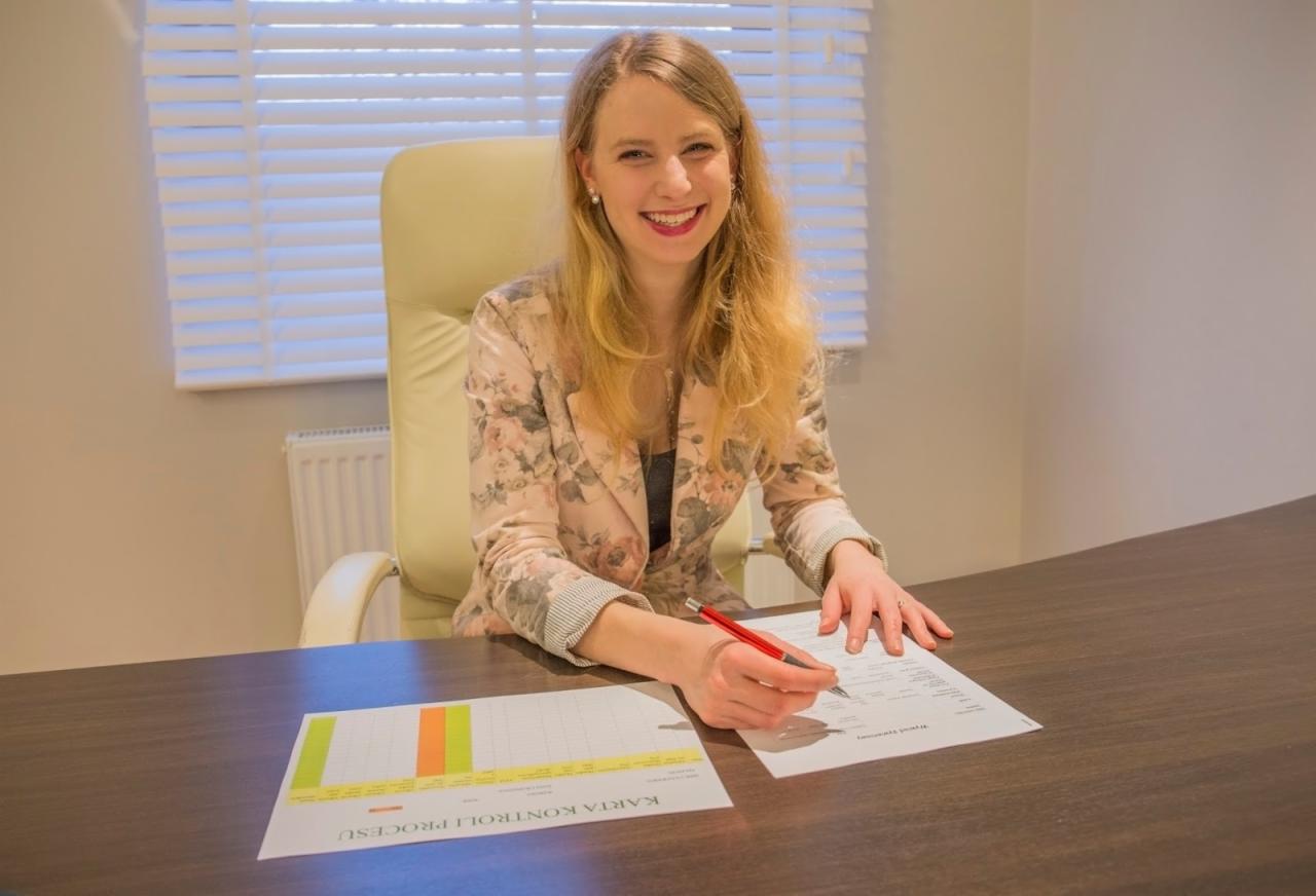 Uśmiechnięta kobieta w kwiecistym żakiecie siedzi przy biurku i wypełnia dokument, obok leży wykres z kolumnami w kolorach żółtym i zielonym.