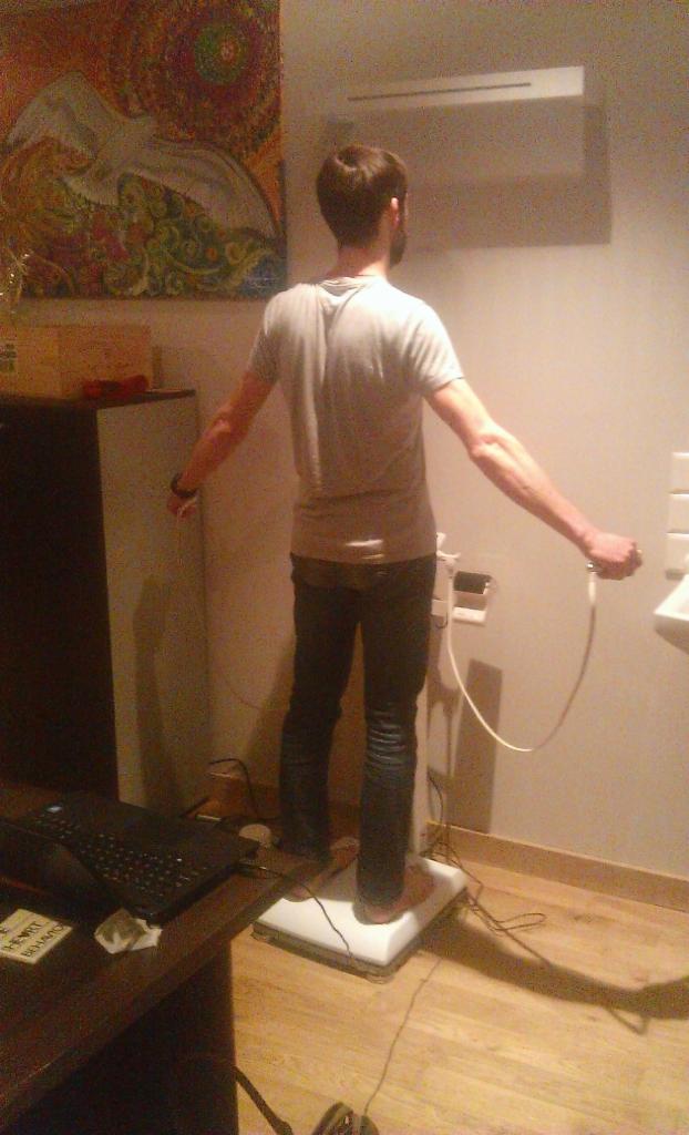 Mężczyzna tyłem, stojący na analizatorze składu ciała, trzymający elektrody w dłoniach, widoczny obraz na ścianie i elementy wyposażenia gabinetu.