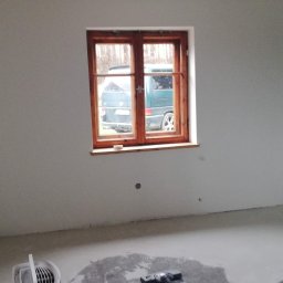 Dom-Zbych - Pokój w trakcie remontu: świeżo położona gładź na ścianach, drewniane okno z widokiem na zewnątrz, narzędzia pracy na parapecie i podłodze. Widoczne ślady prac wykończeniowych.