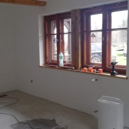 Dom-Zbych - Pokój po gładziowaniu ścian, z drewnianymi oknami i narzędziami na parapecie. Widoczny poziomica na podłodze i biały nawilżacz powietrza. Ściany pomalowane na biało.