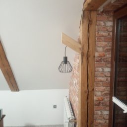 Dom-Zbych - Wykończone wnętrze poddasza z drewnianymi belkami, ceglaną ścianą i nowoczesną lampą wiszącą. Widoczny fragment grzejnika i okna.