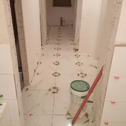 Układanie białych płytek podłogowych z marmurowym wzorem w korytarzu, widoczne plastikowe krzyżyki i poziomica, wiadro z klejem w tle.