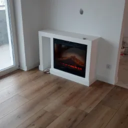 Nowoczesny salon z panelami podłogowymi w odcieniach brązu, białym elektrycznym kominkiem i wyjściem na balkon, widoczne puszki z farbą w przejściu do innego pomieszczenia.