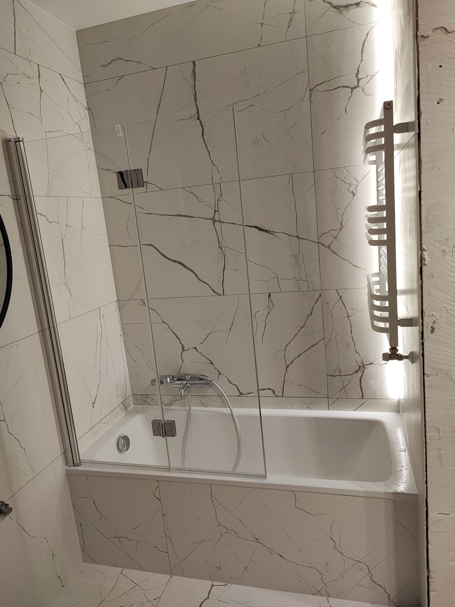 Biała wanna z parawanem nawannowym i grzejnikiem drabinkowym na tle płytek imitujących marmur. Nowoczesna łazienka po remoncie, oświetlenie LED.