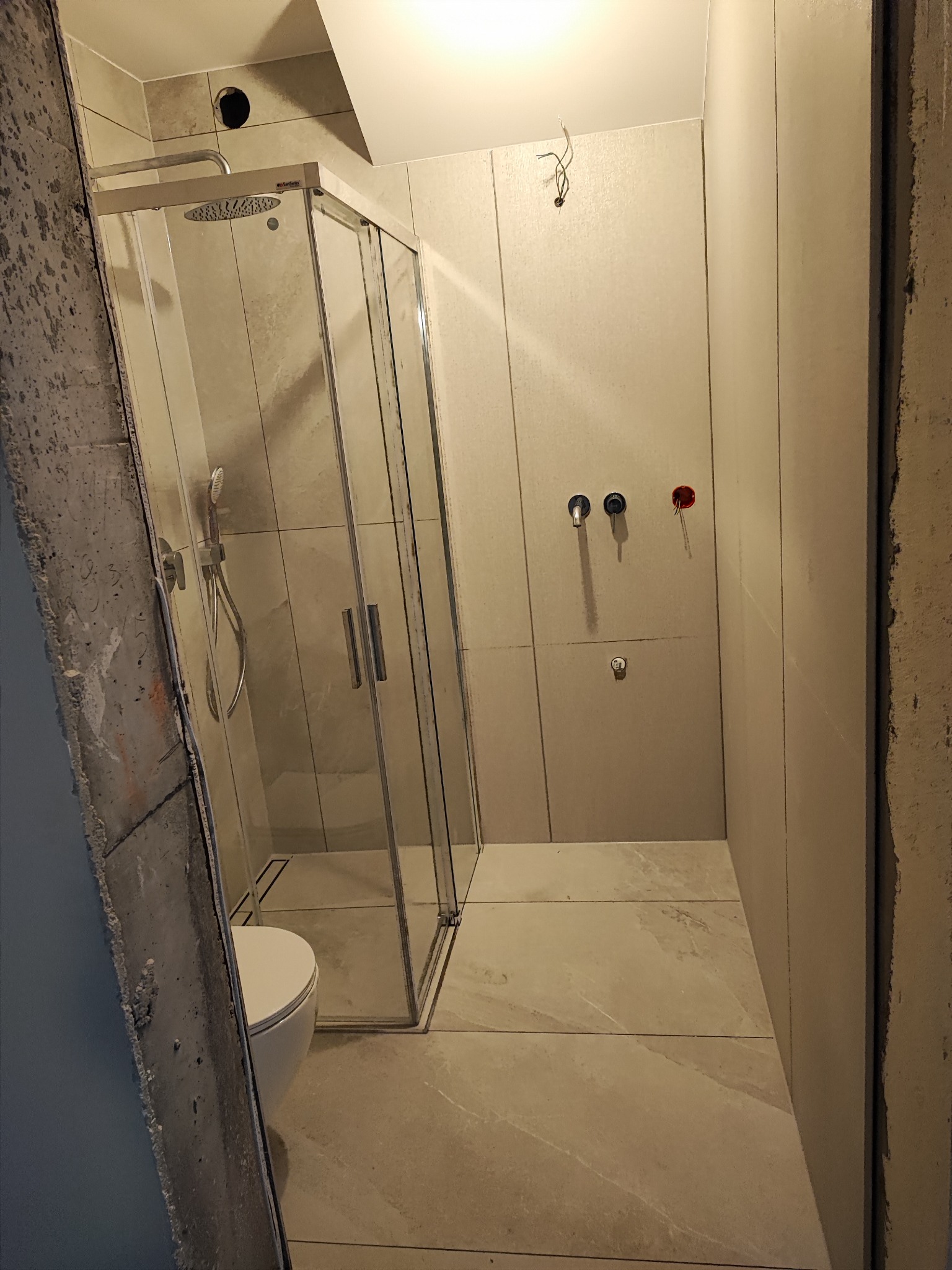 Jasna łazienka w trakcie wykańczania: kabina prysznicowa ze szklanymi drzwiami, biała toaleta, płytki podłogowe imitujące kamień, surowe ściany z instalacją wodną.