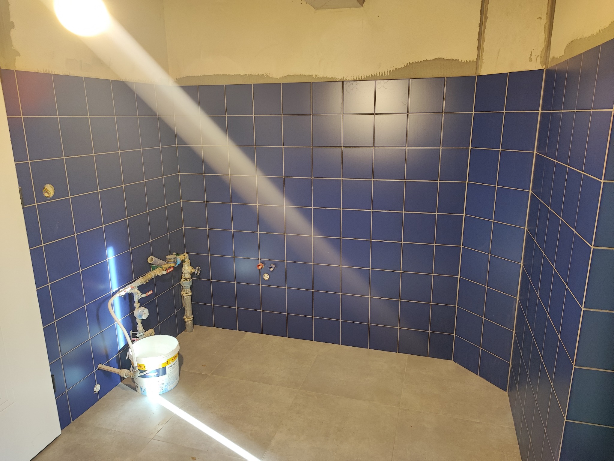 Niebieskie płytki w łazience w trakcie remontu, widoczne instalacje wodne i wiadro z zaprawą. Promień światła podkreśla etap prac wykończeniowych.