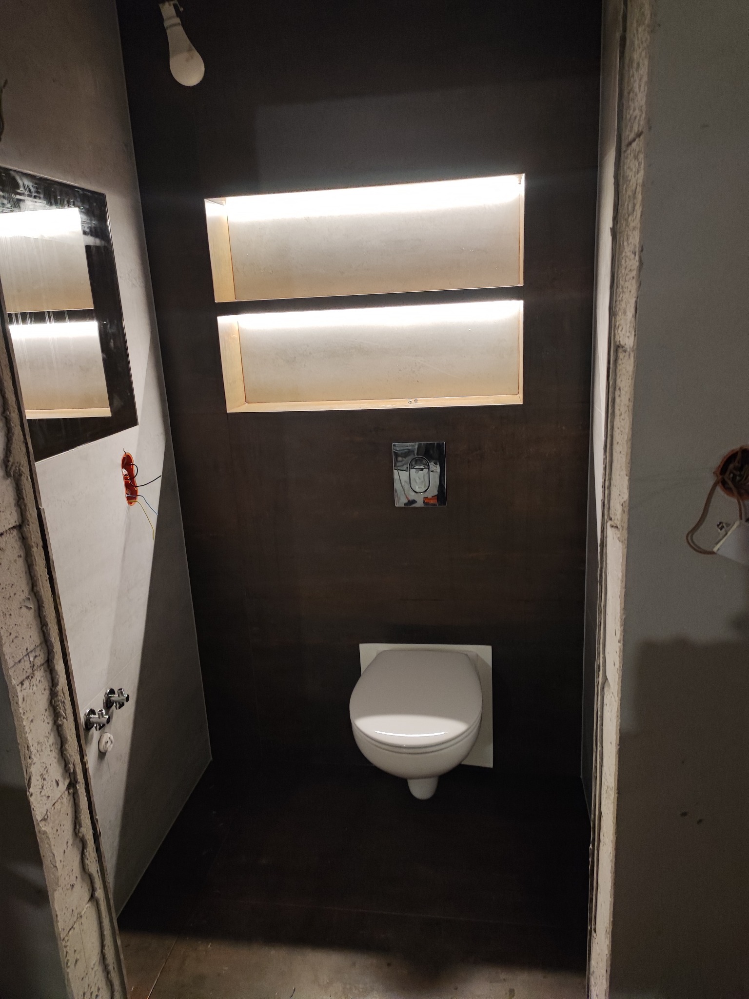 Ciemna łazienka w trakcie wykańczania: podświetlane wnęki, wisząca toaleta, surowe ściany. Widoczne niedokończone instalacje elektryczne i brakujące elementy wykończeniowe.